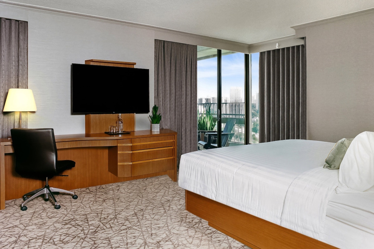 A hotel room with a bed, a desk with a chair, a TV, and a view of the city.