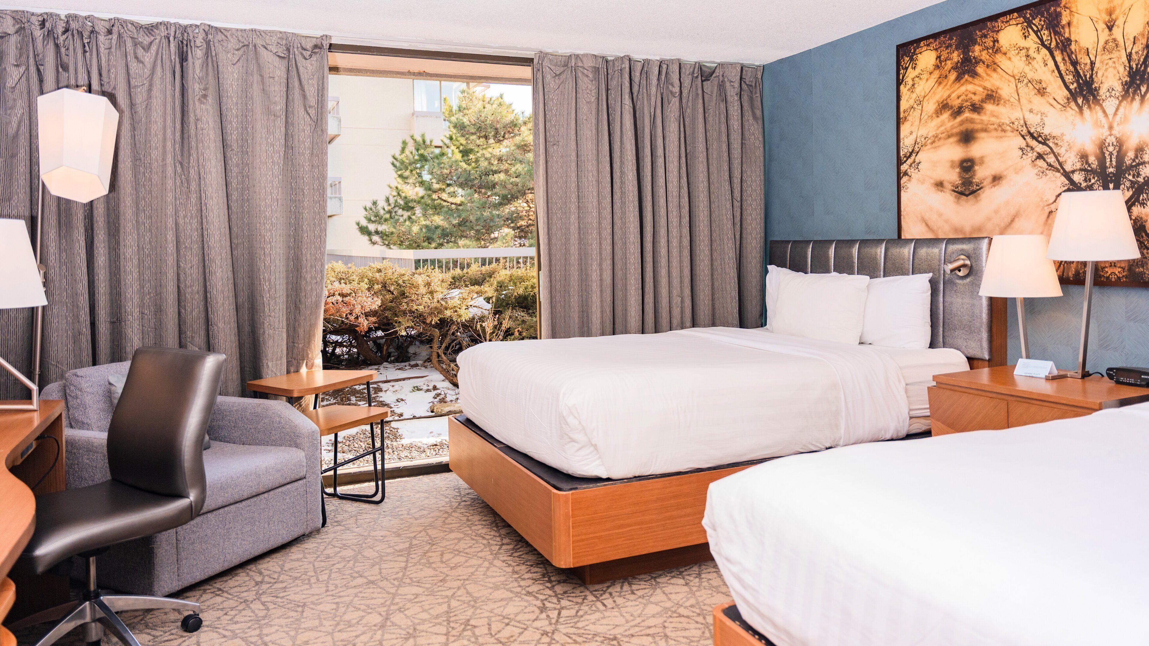 Habitación de hotel con cama, escritorio, silla, mesita de noche y una ventana grande con cortinas.