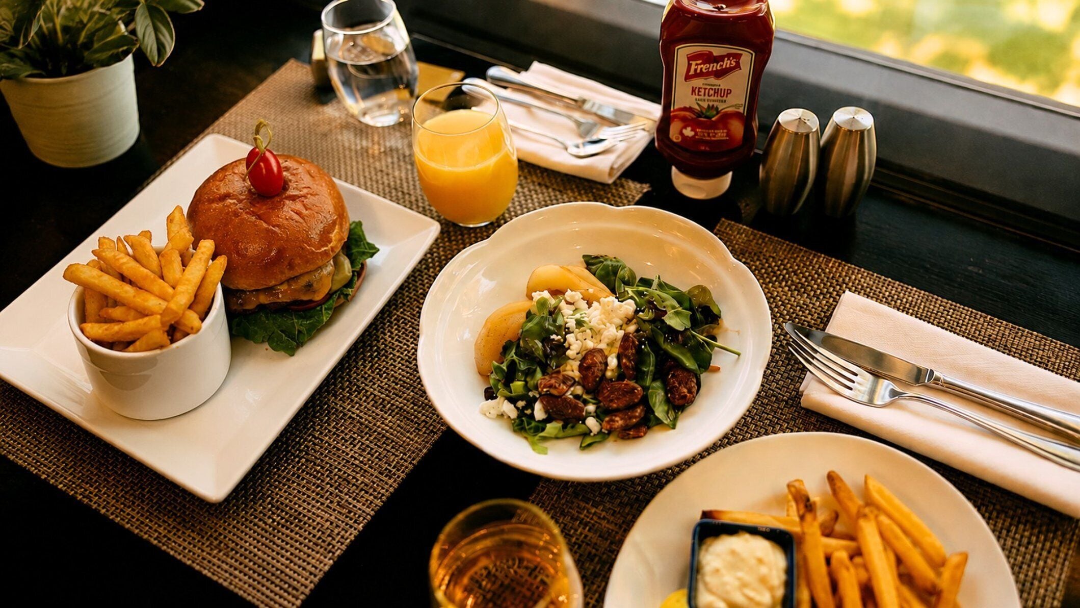 Eine Mahlzeit mit einem Burger, Pommes, Salat und einem Glas Orangensaft auf einem Tisch mit Blick.
