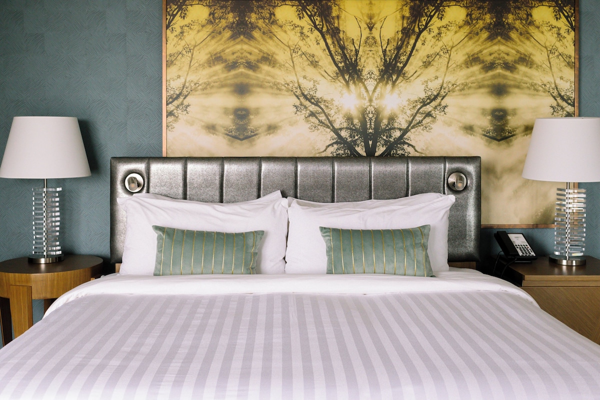 A neatly made bed with striped bedding, two bedside tables with lamps, and a large tree-themed painting on the wall.