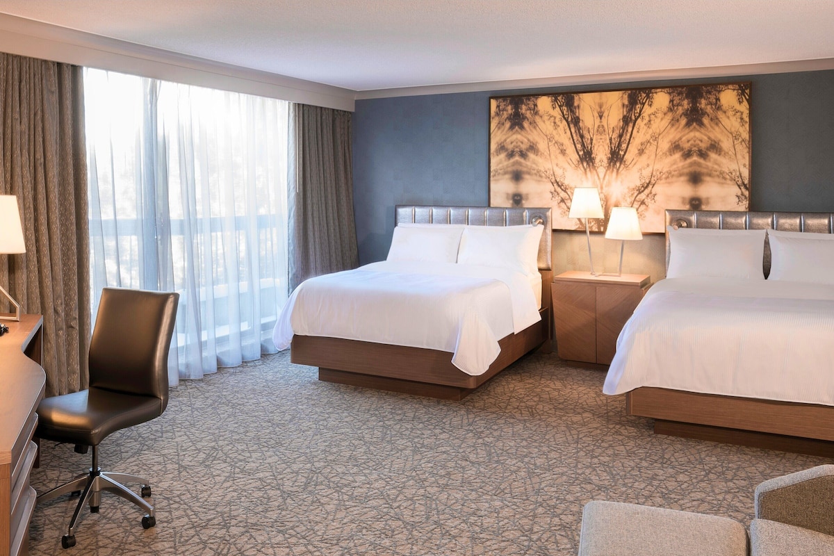 A hotel room with two beds, a desk, a chair, and a large window with curtains.