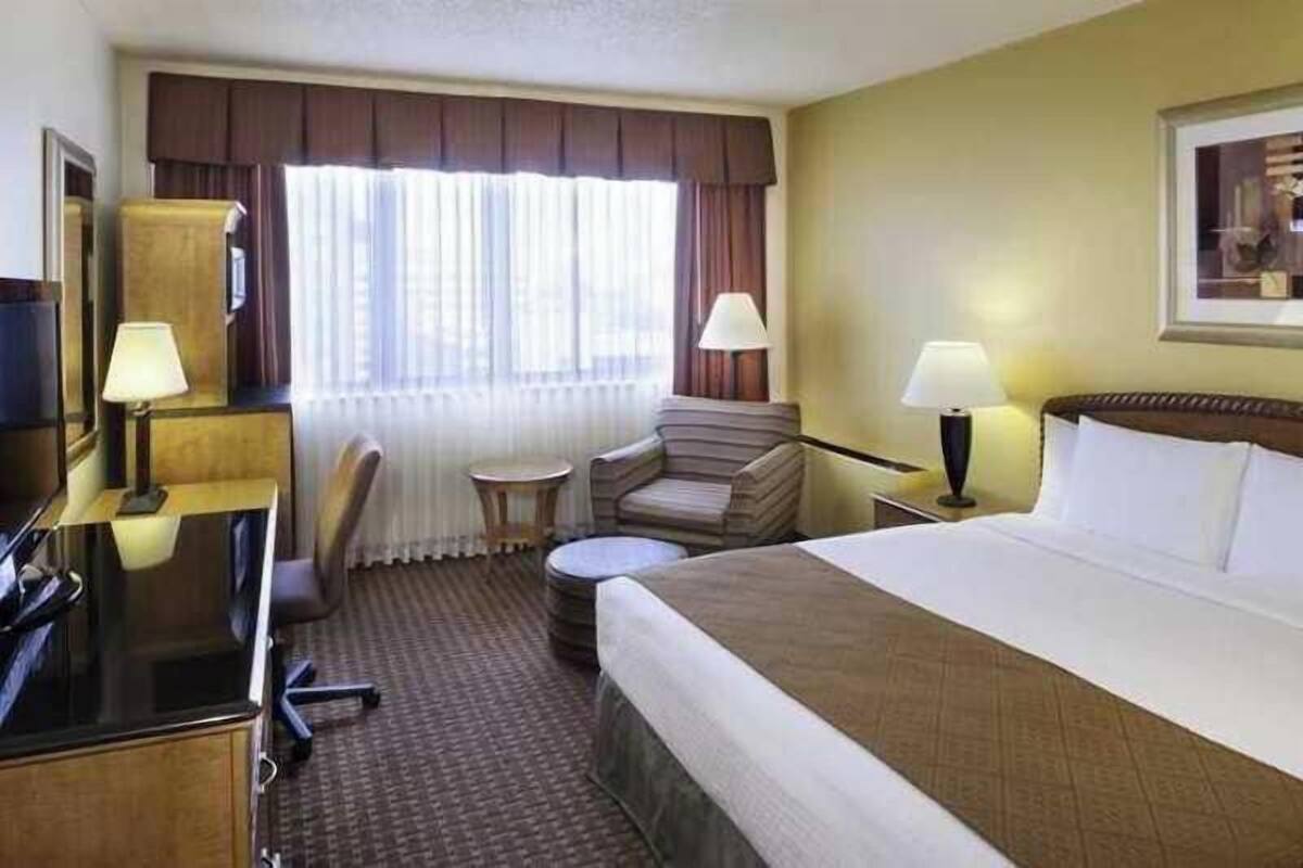 Quarto de hotel com cama, escrivaninha, cadeira, luminária e janela com cortinas.
