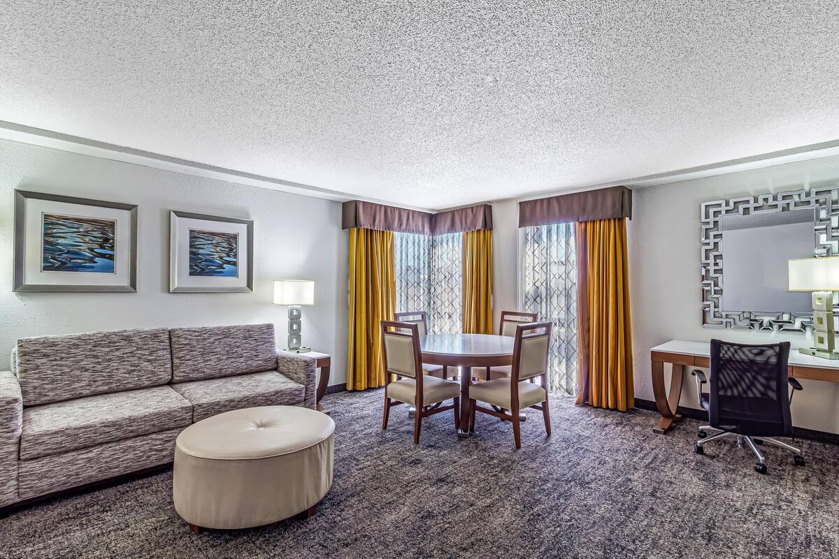 A hotel room with a sofa, ottoman, dining table with chairs, a desk with a chair, a mirror, and two framed artworks on the wall.