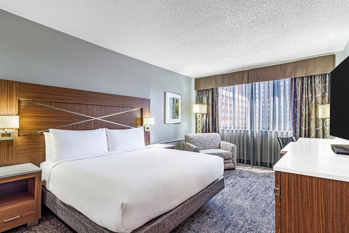 A hotel room with a large bed, a desk, a chair, and a view of the city through the window.