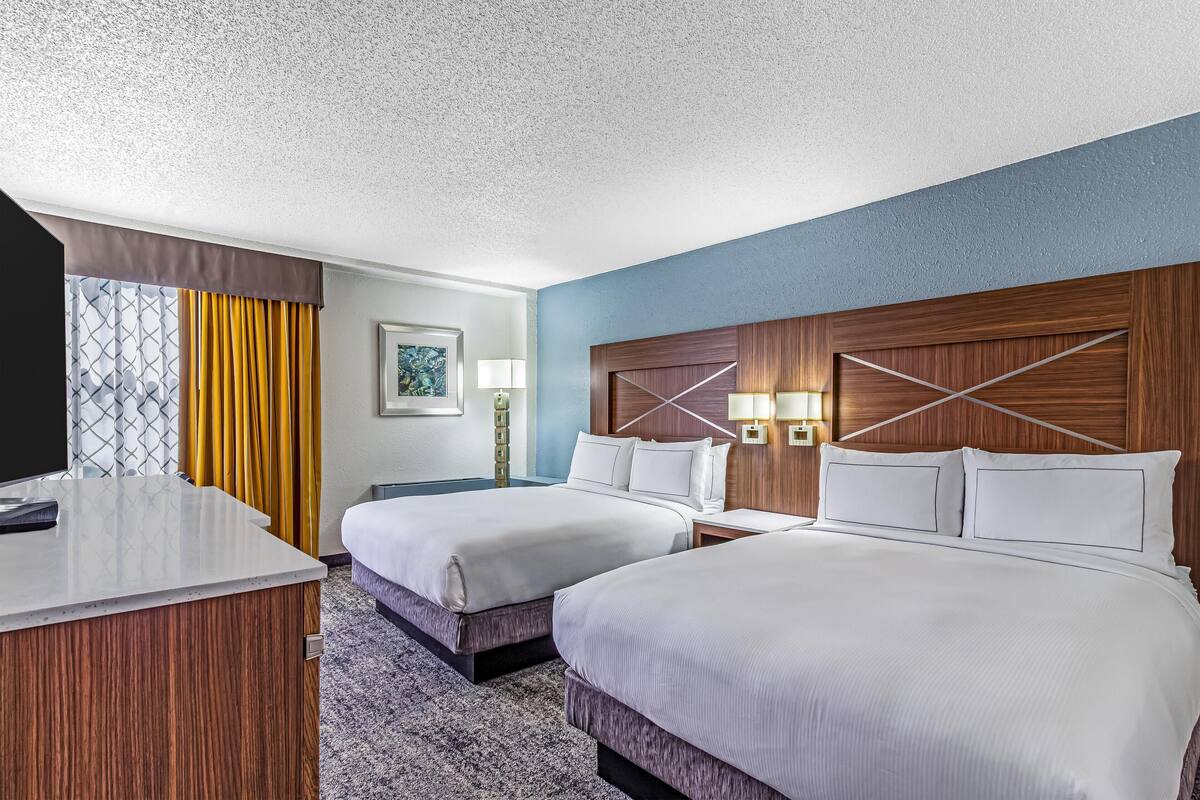 A hotel room with two beds, a desk, a nightstand, a lamp, and a window with curtains.
