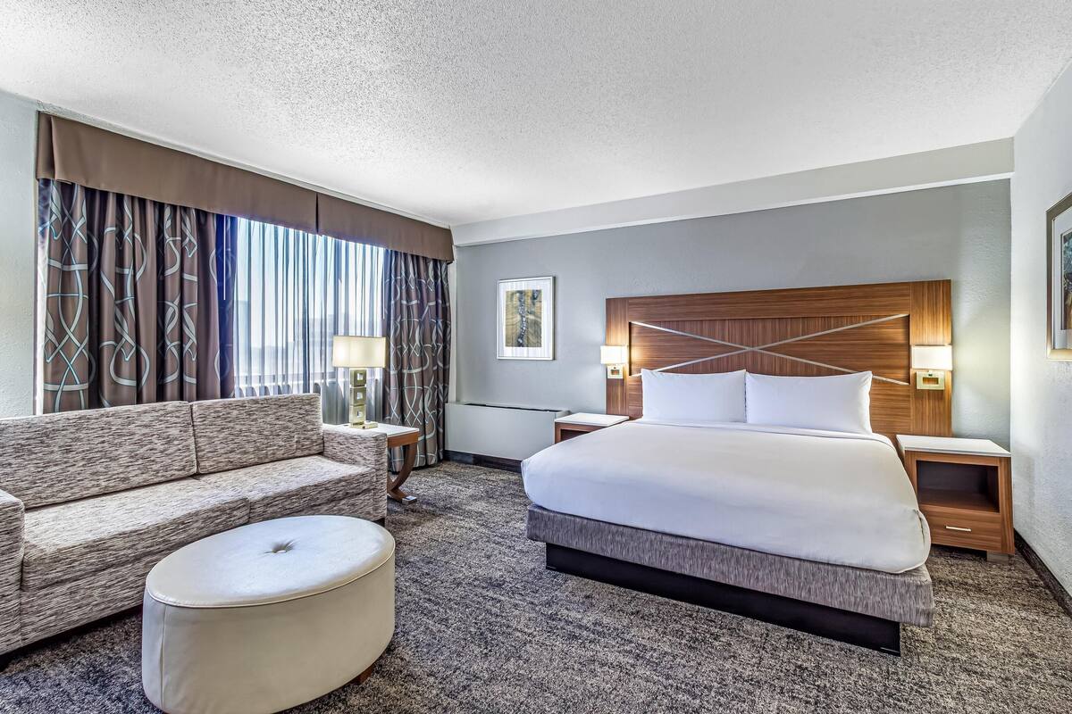 A hotel room with a large bed, a sofa, a round ottoman, a nightstand, a lamp, and a framed picture on the wall.