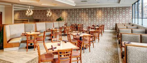 Comedor de hotel con cabinas y mesas, alfombra estampada e iluminación colgante.