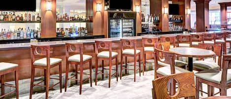 Zona de bar con sillas y mesas de madera, un refrigerador y estantes con botellas.