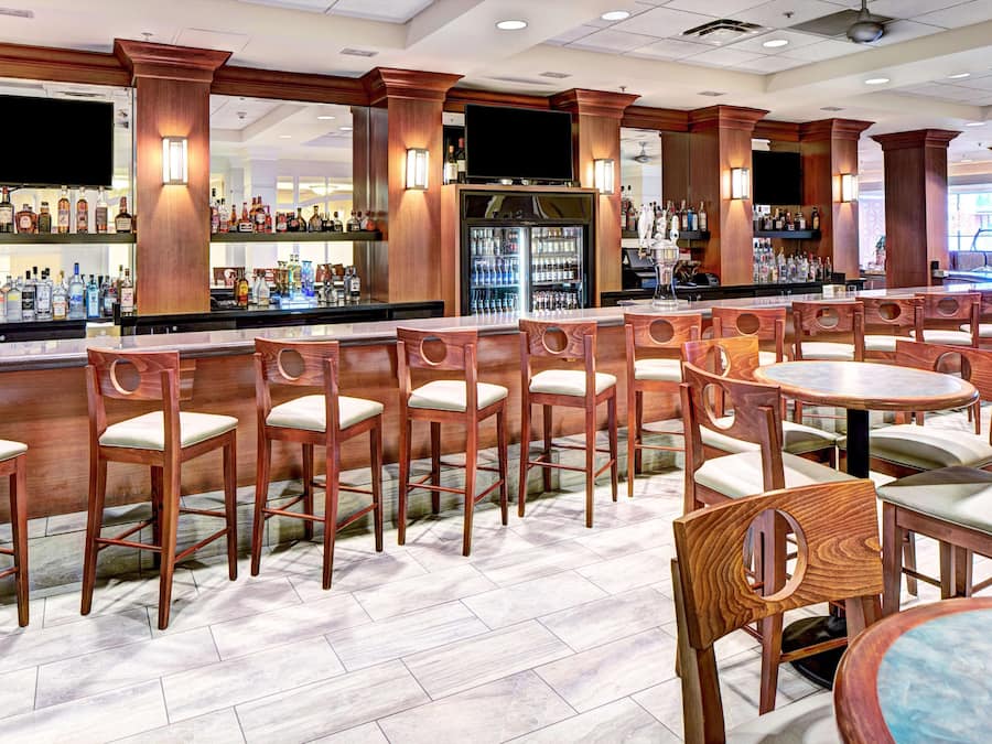 A bar area with wooden chairs and tables, a refrigerator, and shelves with bottles.