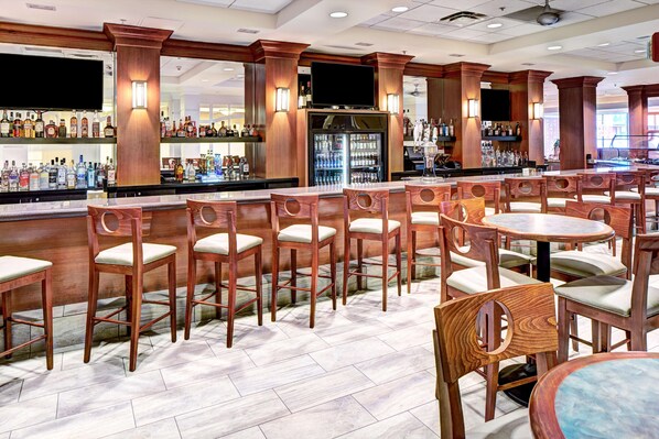 Zona de bar con sillas y mesas de madera, un refrigerador y estantes con botellas.
