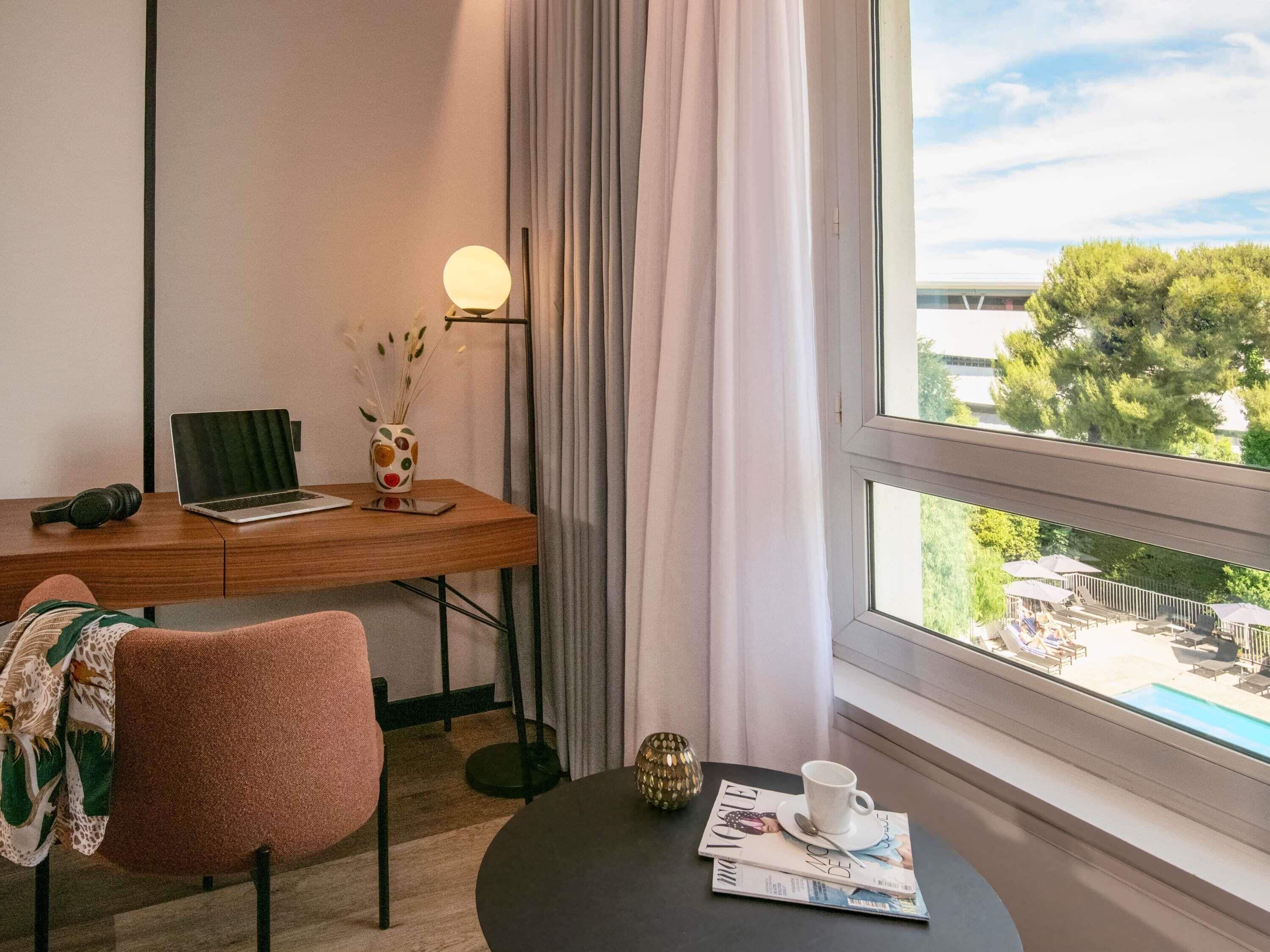 Habitación de hotel con escritorio, una laptop, una silla, una mesita con una revista, una taza y vistas a un espacio exterior con sombrillas.