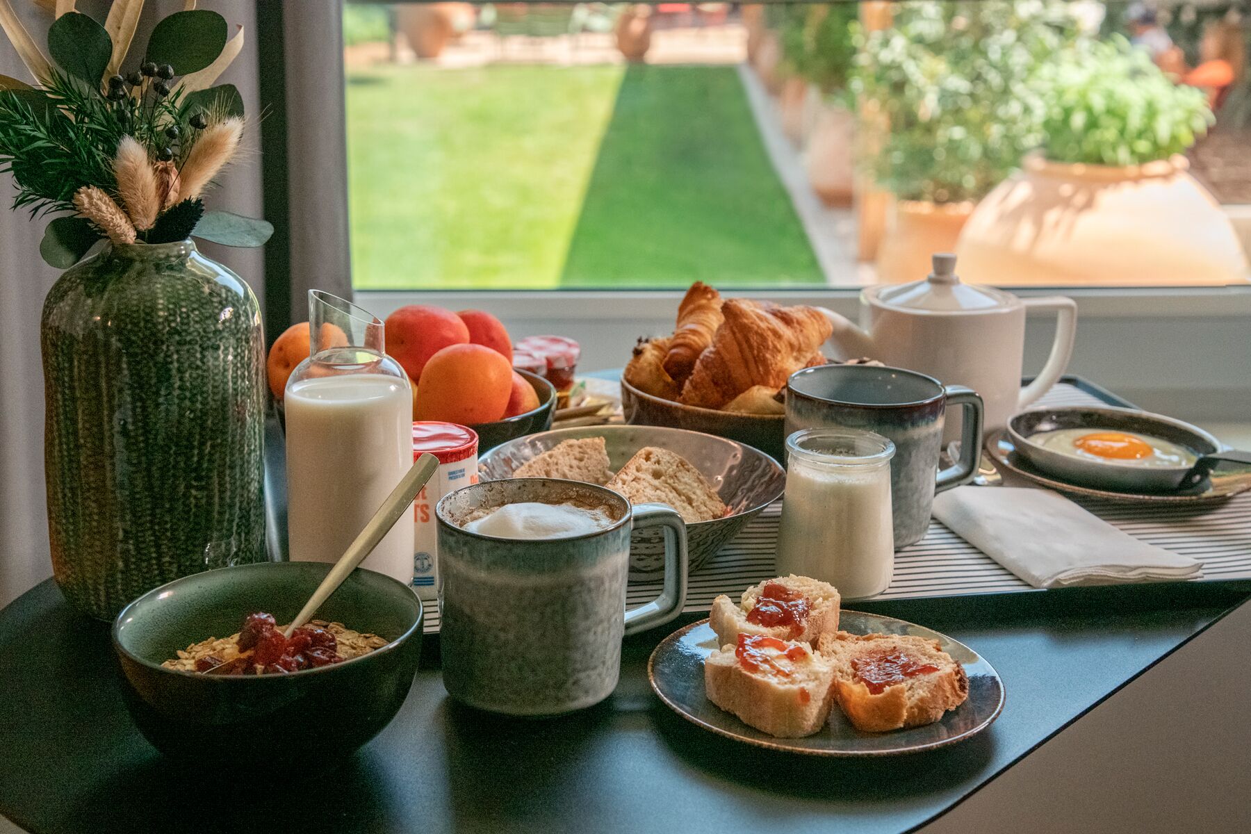 A breakfast table with various food items, a vase with dried plants, and a view of a garden through a window.