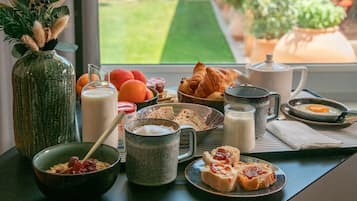 A breakfast table with various food items, a vase with dried plants, and a view of a garden through a window.