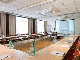 A conference room with a long table, chairs, water bottles, and a presentation screen displaying "NOVOVETEL HOTELS".