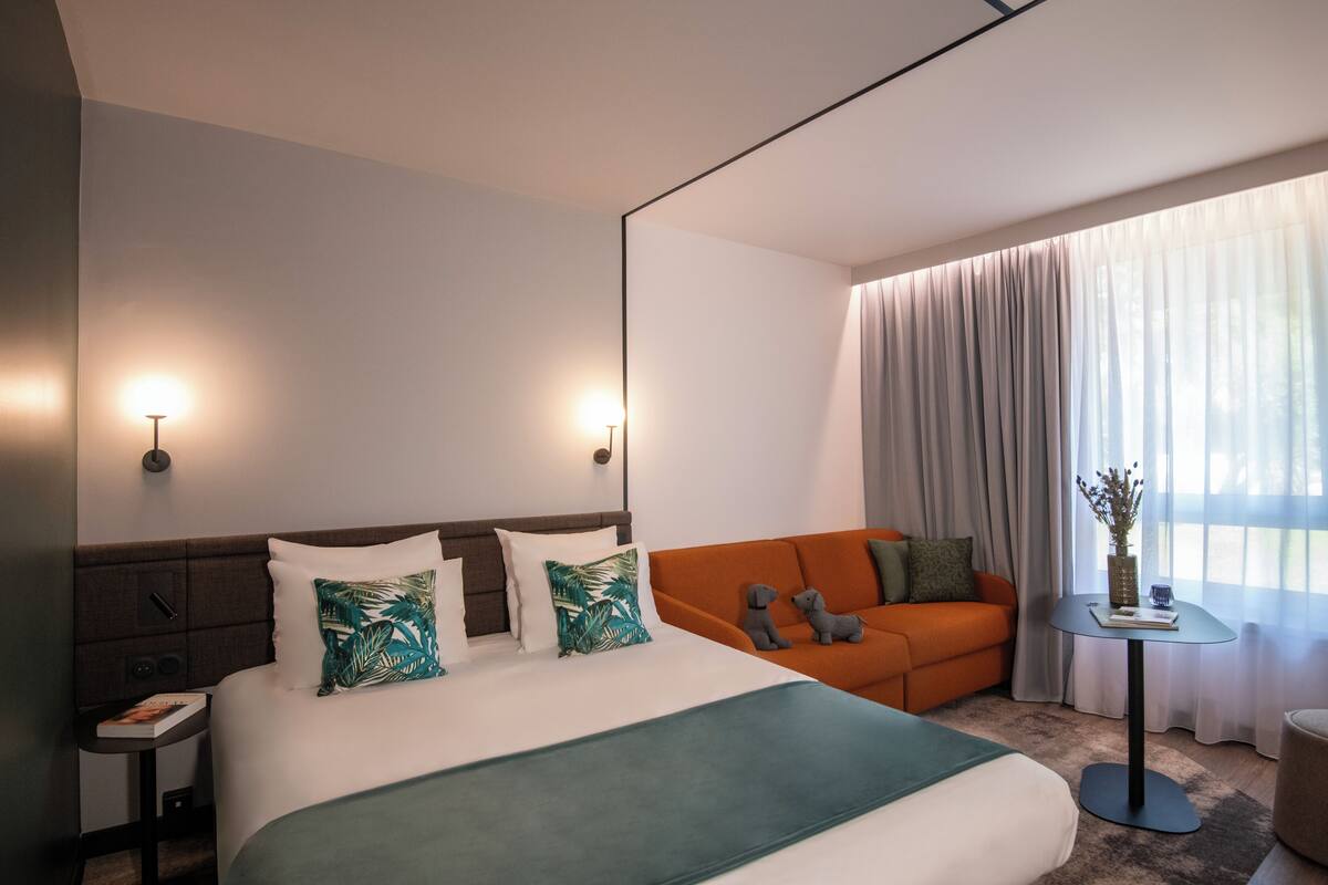 A hotel room with a large bed, an orange sofa, a small table with a vase, and a nightstand with a lamp.