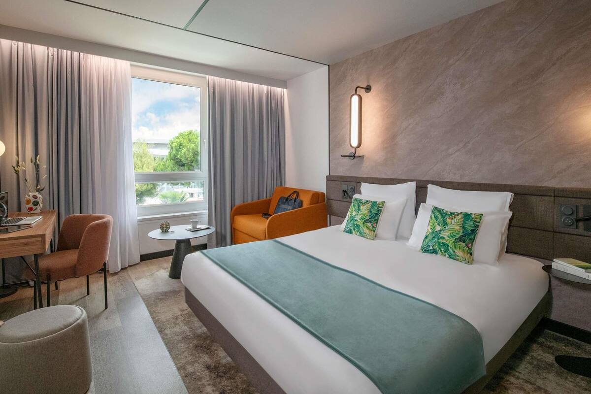 A hotel room with a bed, a desk, a chair, a small table, and a window with curtains.