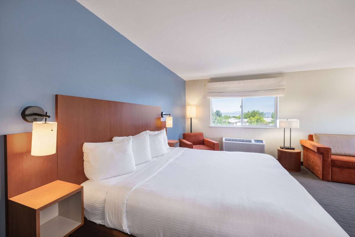 A hotel room with a large bed, two bedside tables with lamps, a chair, a small table, and a window with a view.