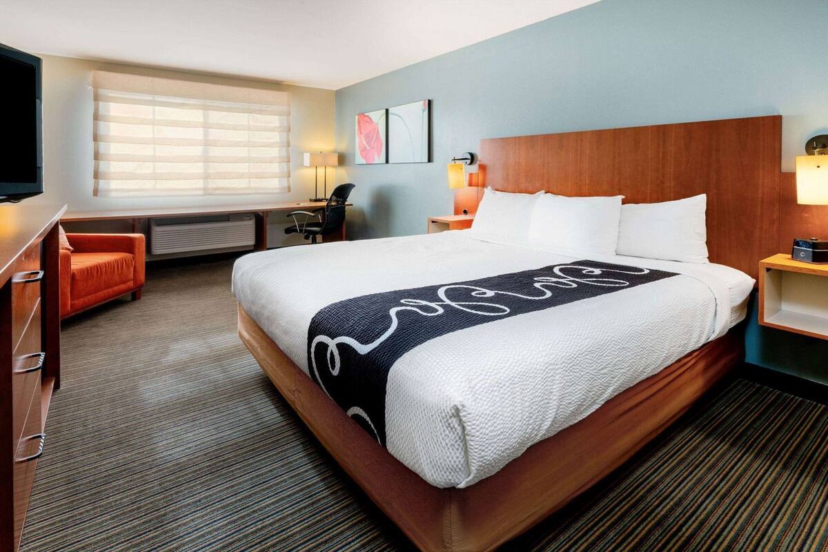 A hotel room with a large bed, a desk, a chair, a TV, and a window with blinds.