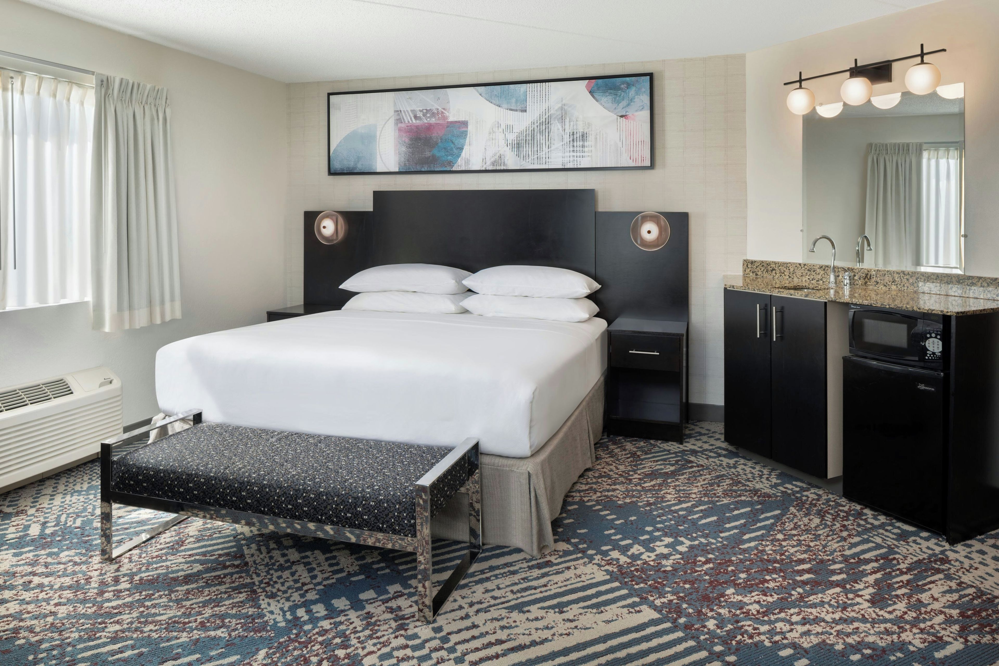 A hotel room with a bed, bedside tables, a bench, a large mirror, and a wall-mounted artwork.