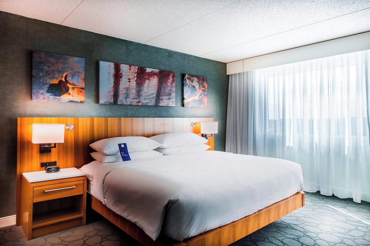 Une chambre d’hôtel avec un grand lit, deux tables de chevet, une petite table et une lampe fixée au mur. Trois œuvres d’art sont accrochées au-dessus du lit.