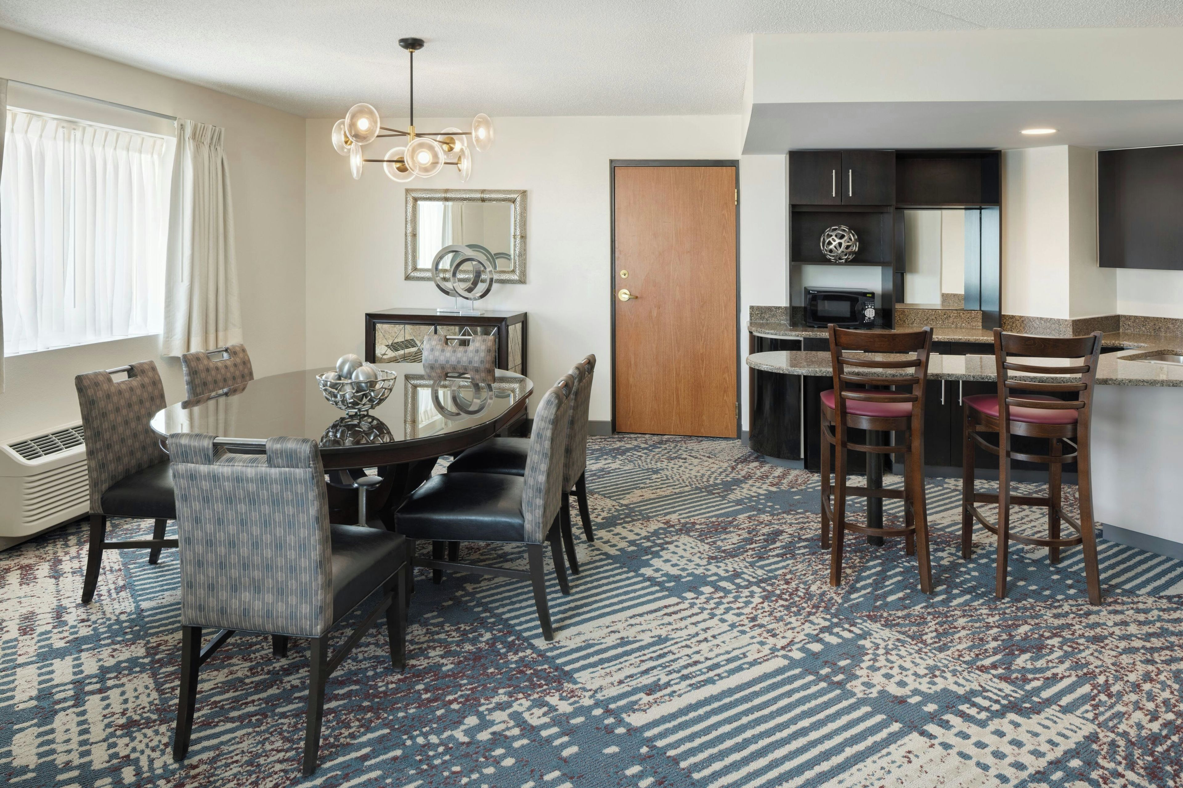 A hotel room with a dining table, chairs, a kitchen area, and a large mirror on the wall.