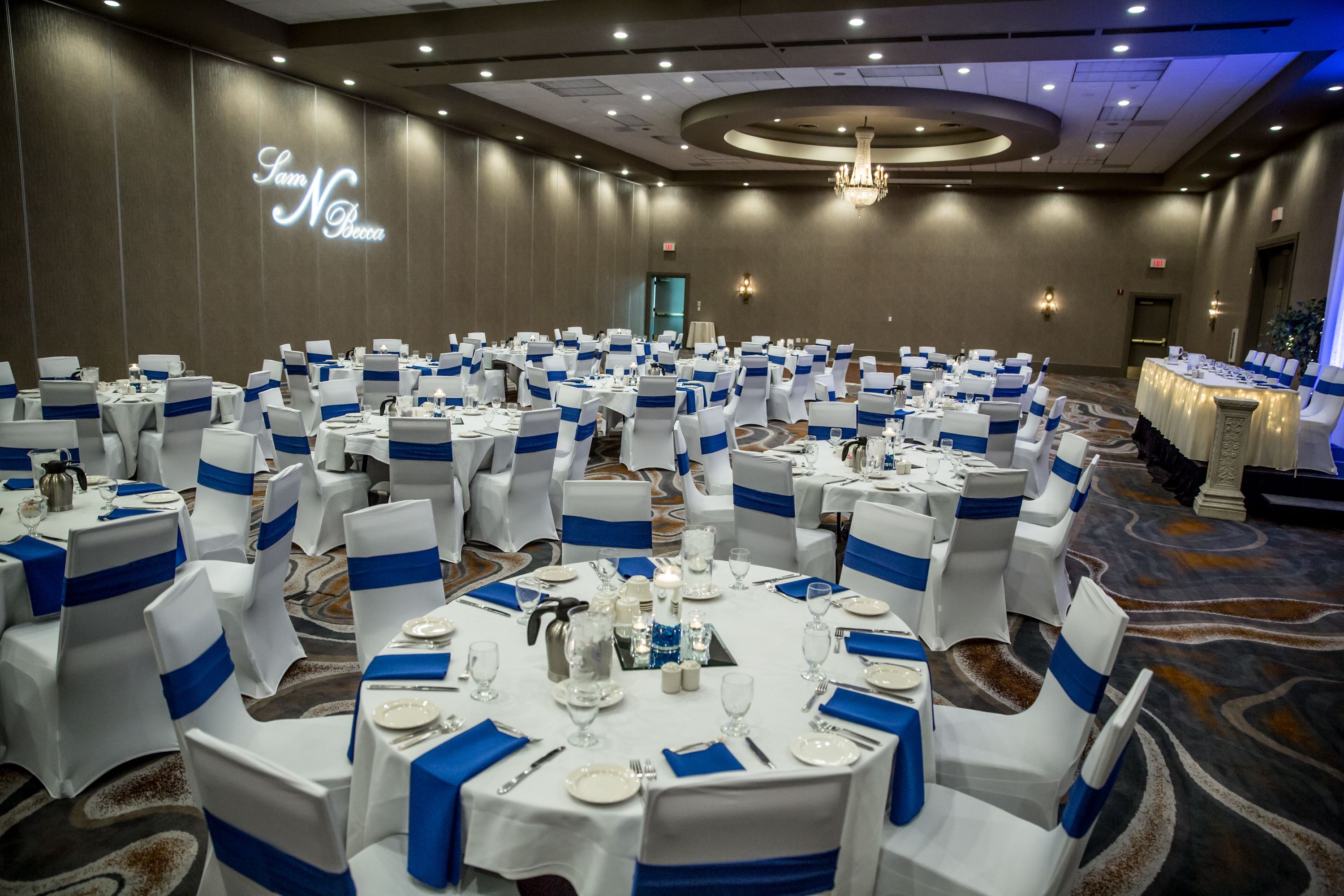 A banquet hall with round tables set for a formal event, featuring blue sashes on chairs and white tablecloths.