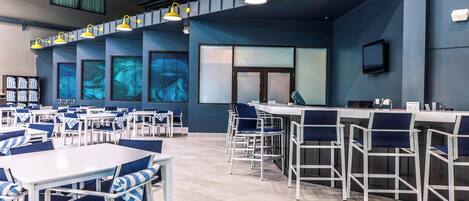 A modern cafeteria with white tables and blue chairs, yellow pendant lights, and large windows with artwork.