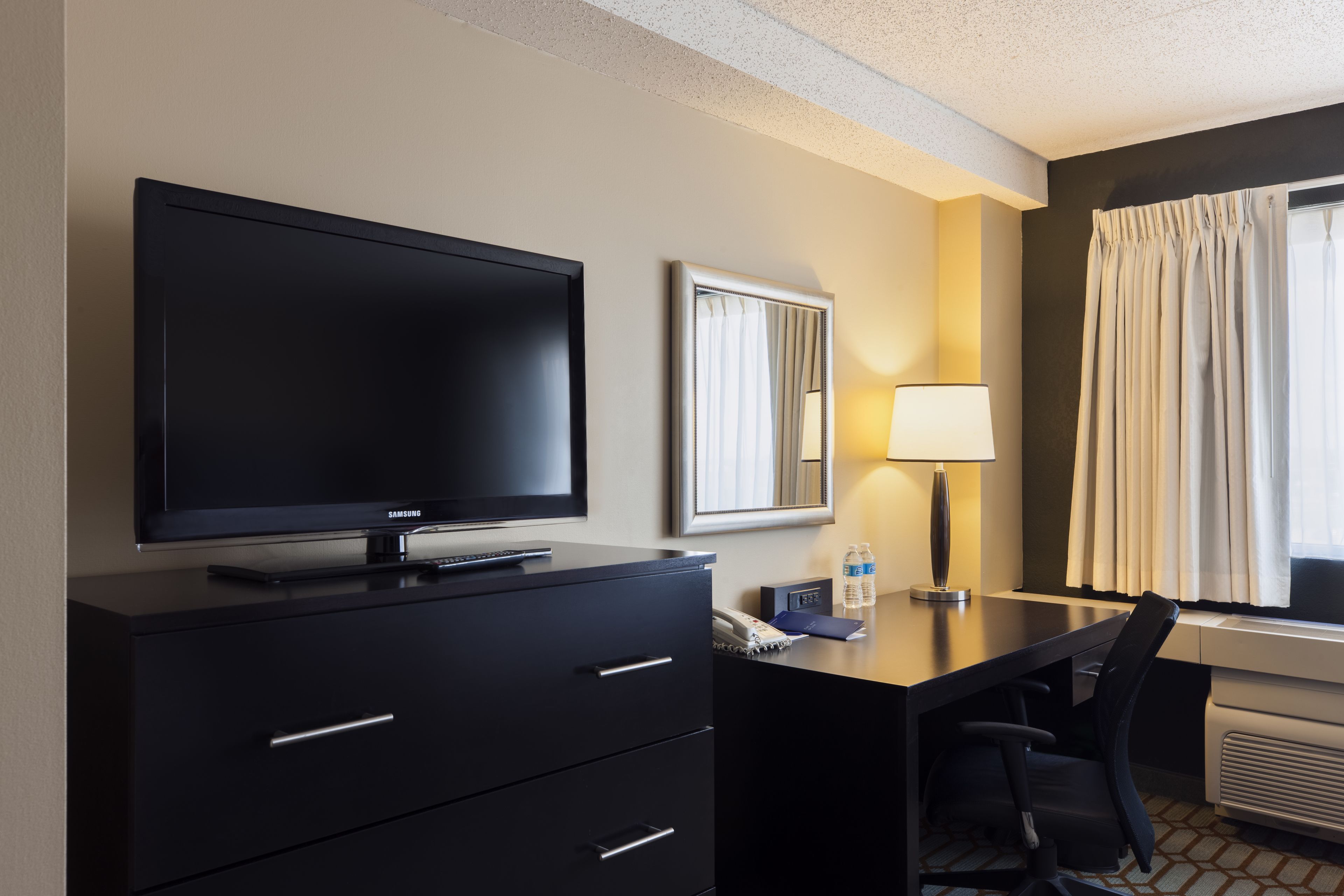 Habitación de hotel con televisión de pantalla plana sobre un mueble negro, un escritorio con silla, una lámpara, un espejo y una ventana con cortinas.