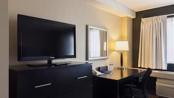 A hotel room with a flat-screen TV on a black cabinet, a desk with a chair, a lamp, a mirror, and a window with curtains.