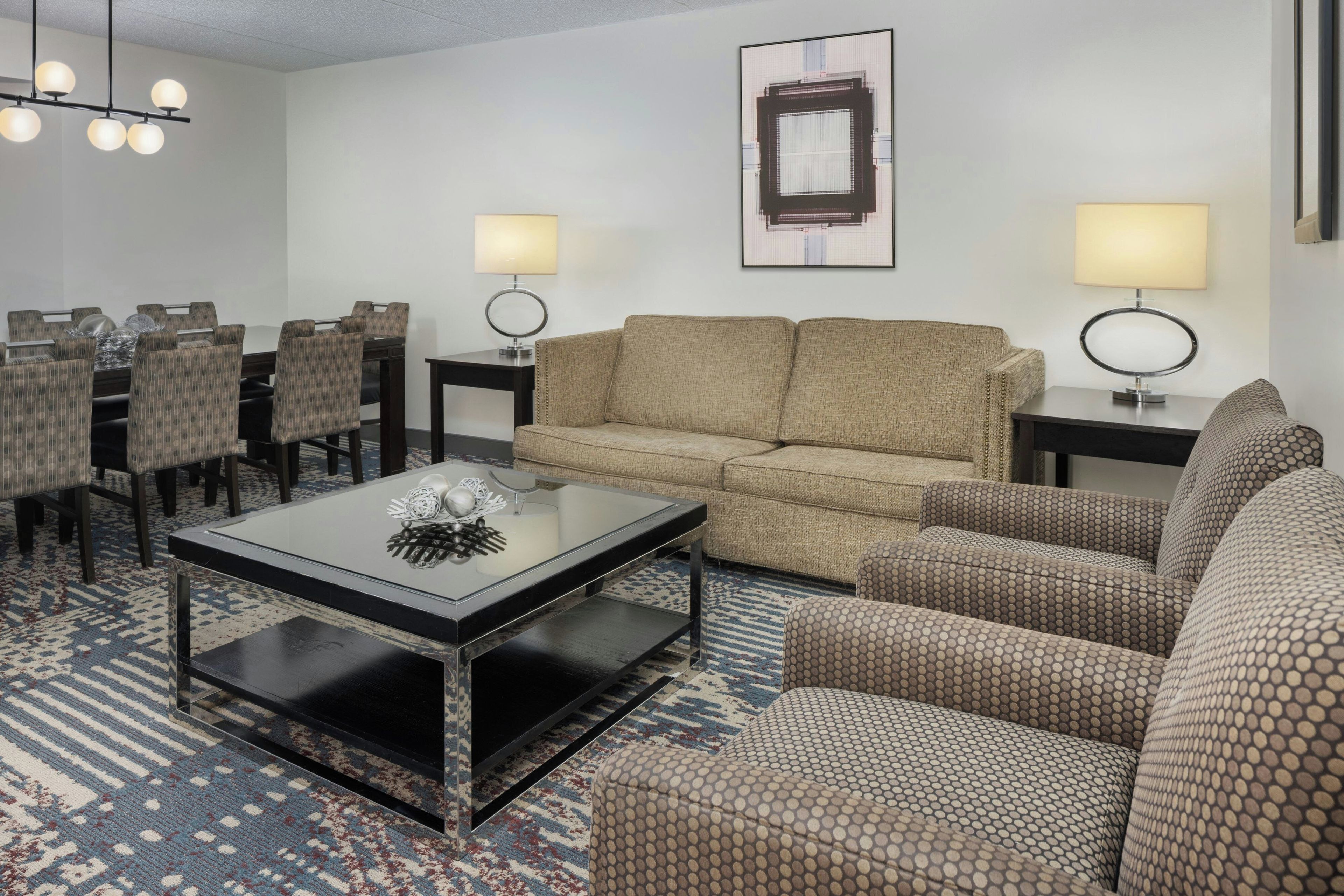 A hotel room with a sofa, armchairs, a coffee table, a dining table with chairs, and two bedside lamps.