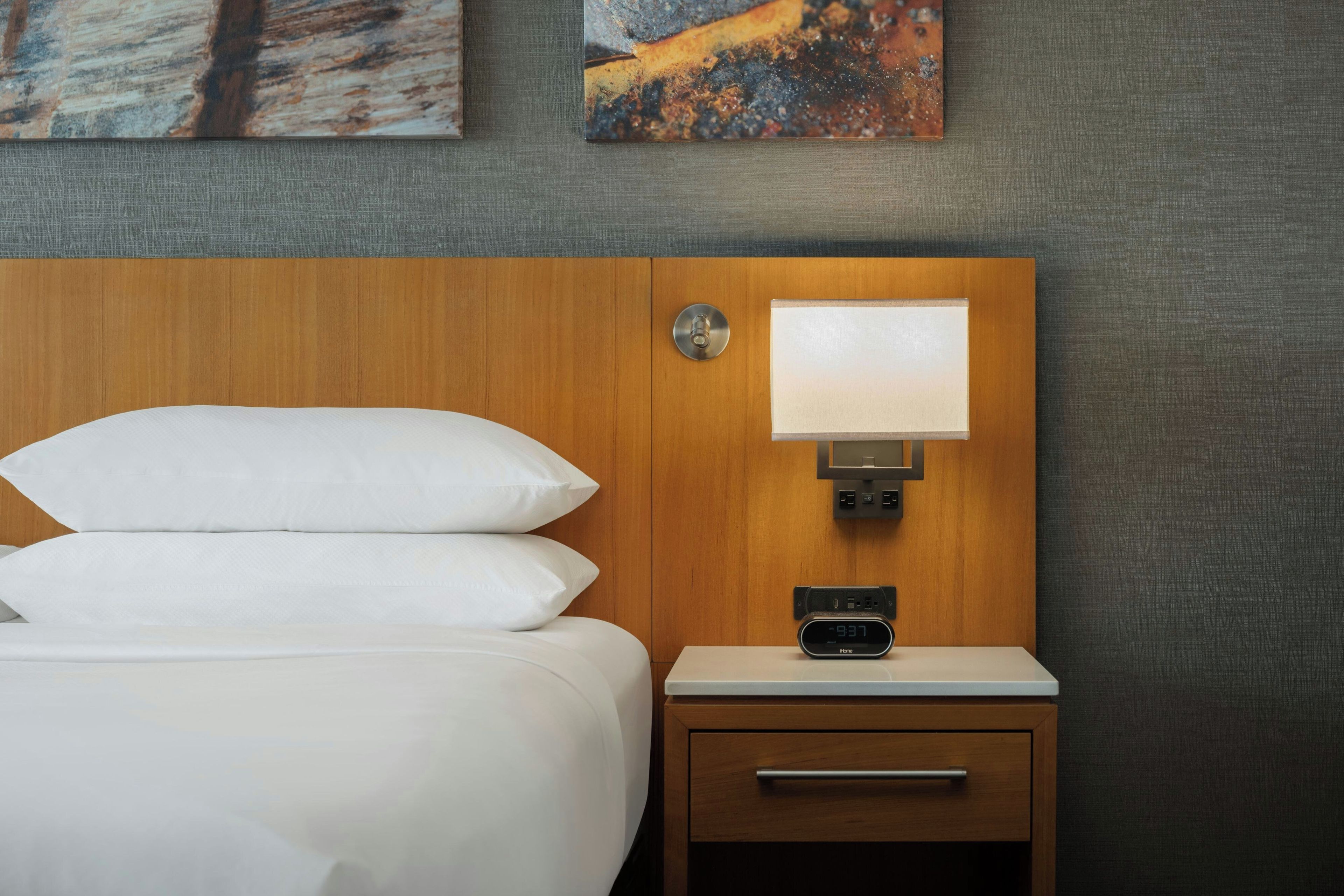 Une chambre d’hôtel avec un lit, une table de chevet avec une lampe, une horloge numérique et des œuvres d’art accrochées au mur.