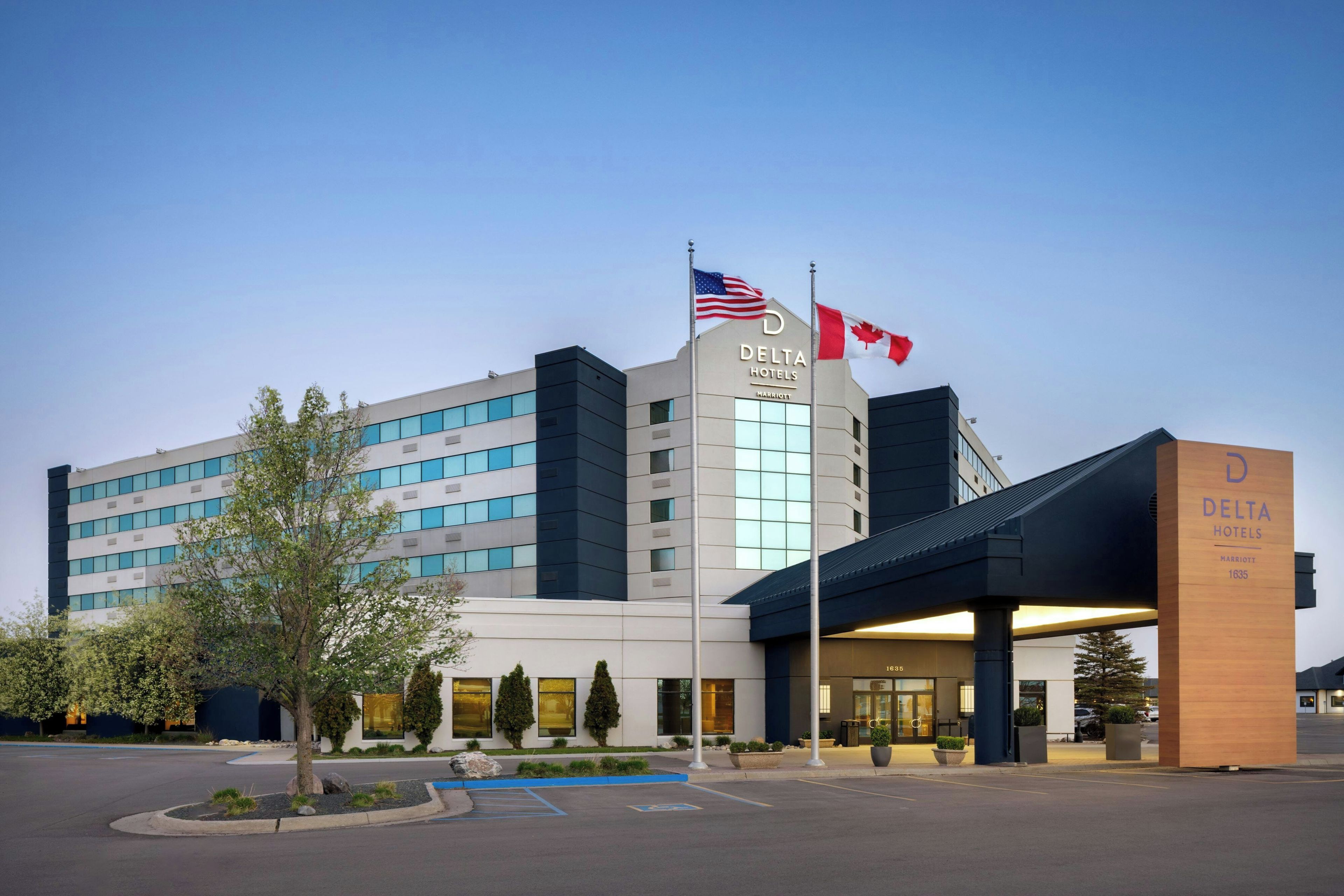 A modern hotel building with a prominent 'Delta Hotels' sign, a flagpole with the American flag, and a flagpole with the Canadian flag.