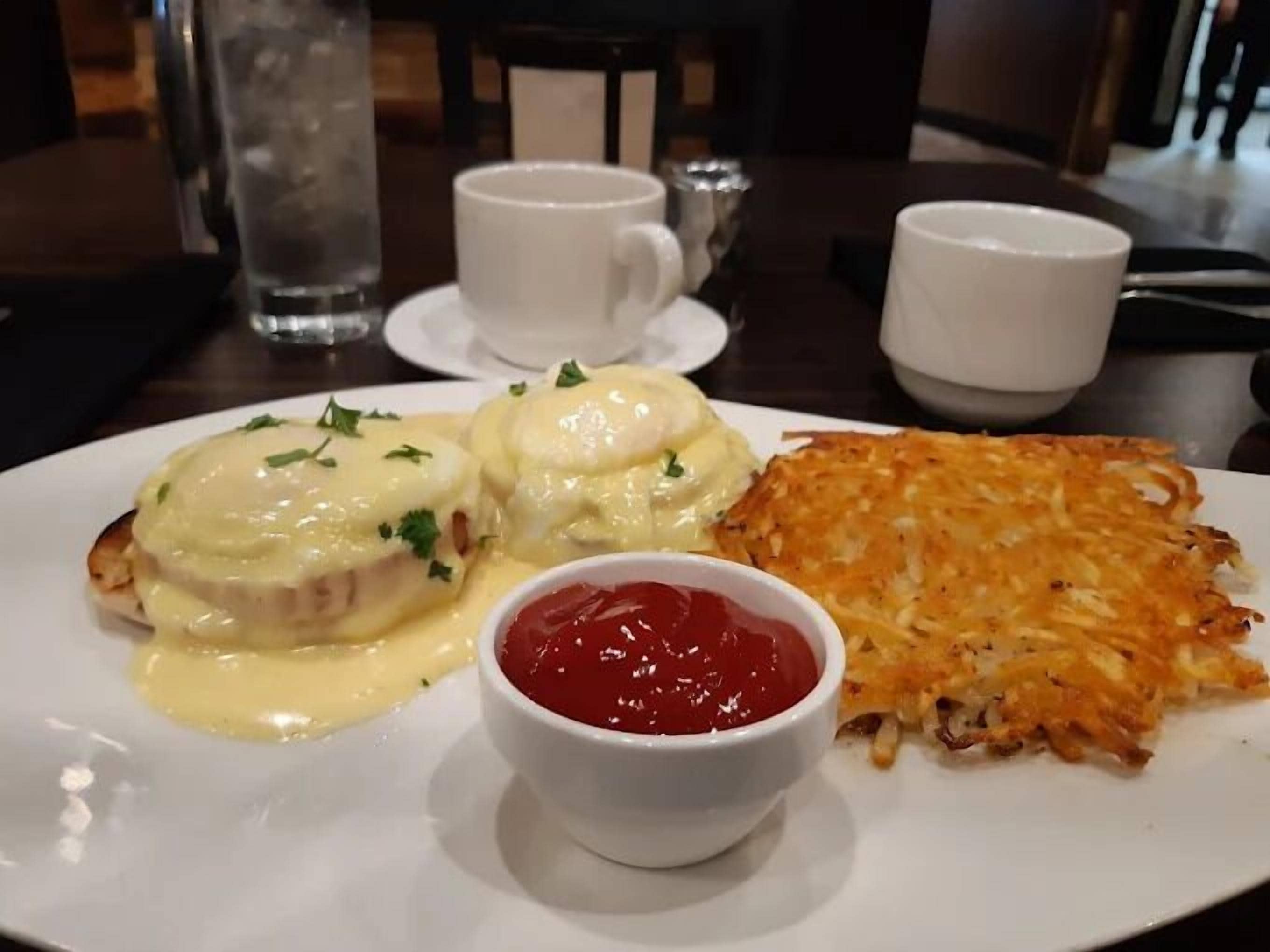 A plate with eggs benedict, hash browns, and ketchup.