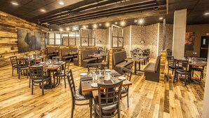 A restaurant with wooden flooring, brick walls, and modern lighting fixtures. Tables and chairs are arranged for dining.