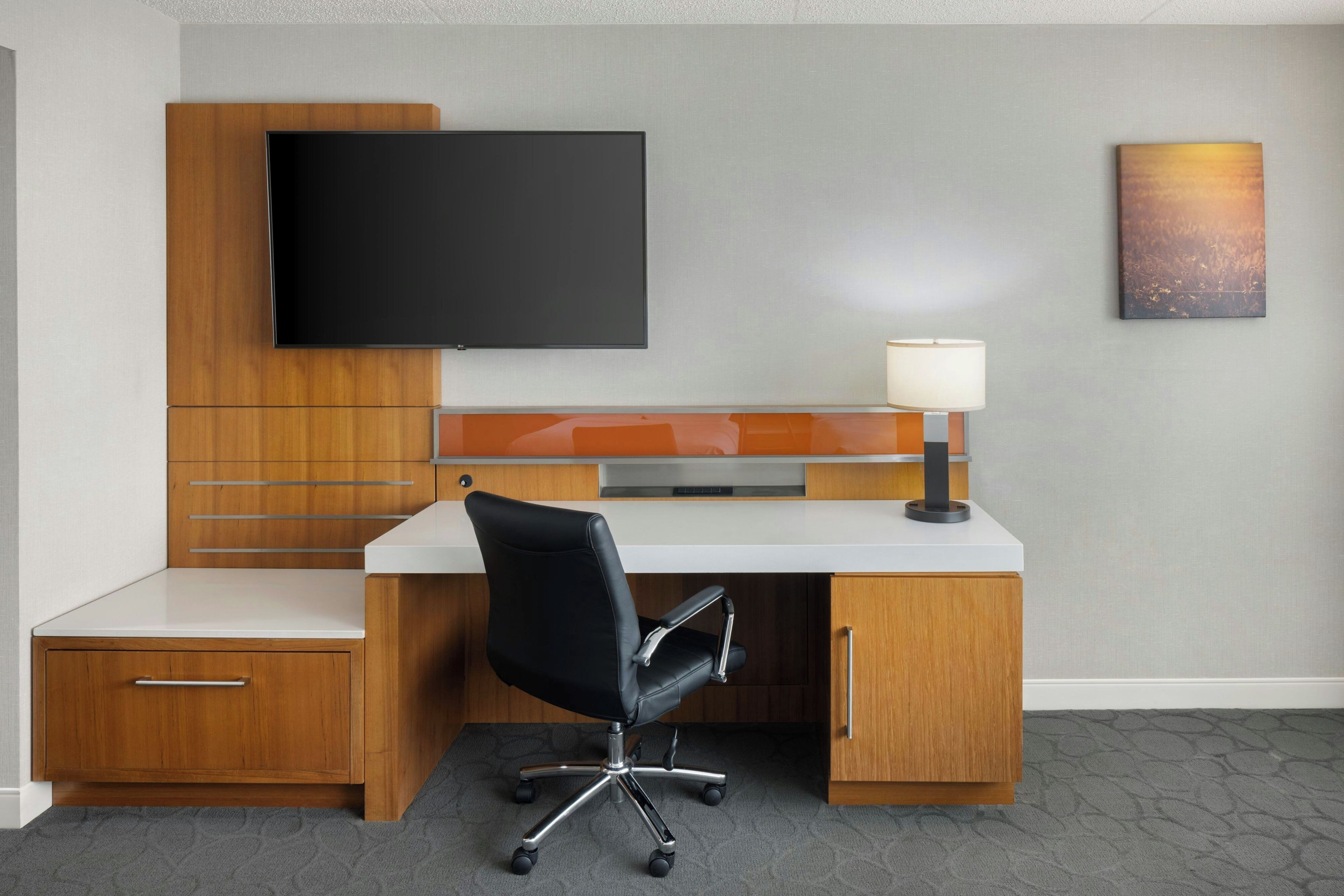 A hotel room with a desk, chair, television, and artwork on the wall.