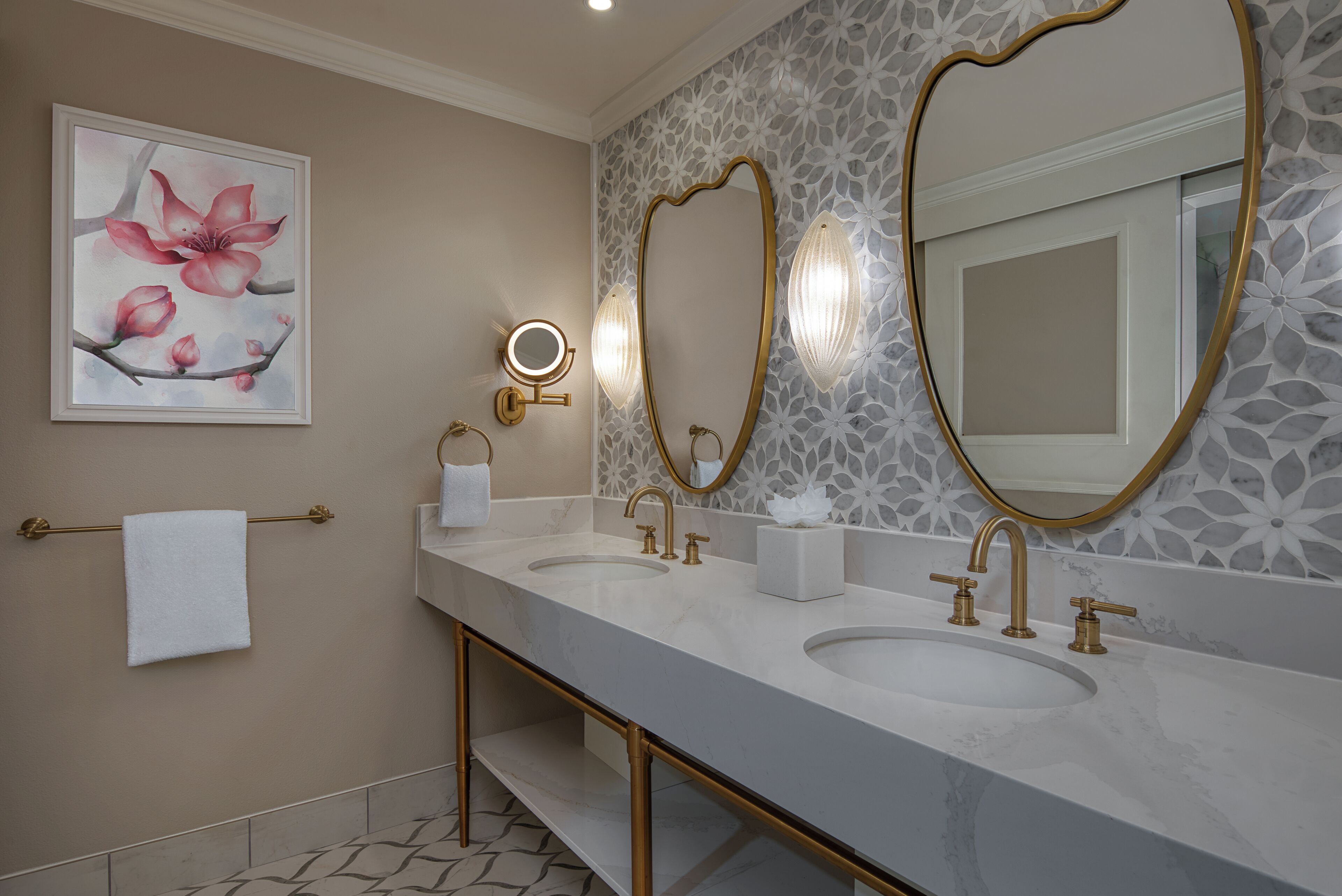 A bathroom with two sinks, gold fixtures, a large mirror, and a floral artwork on the wall.
