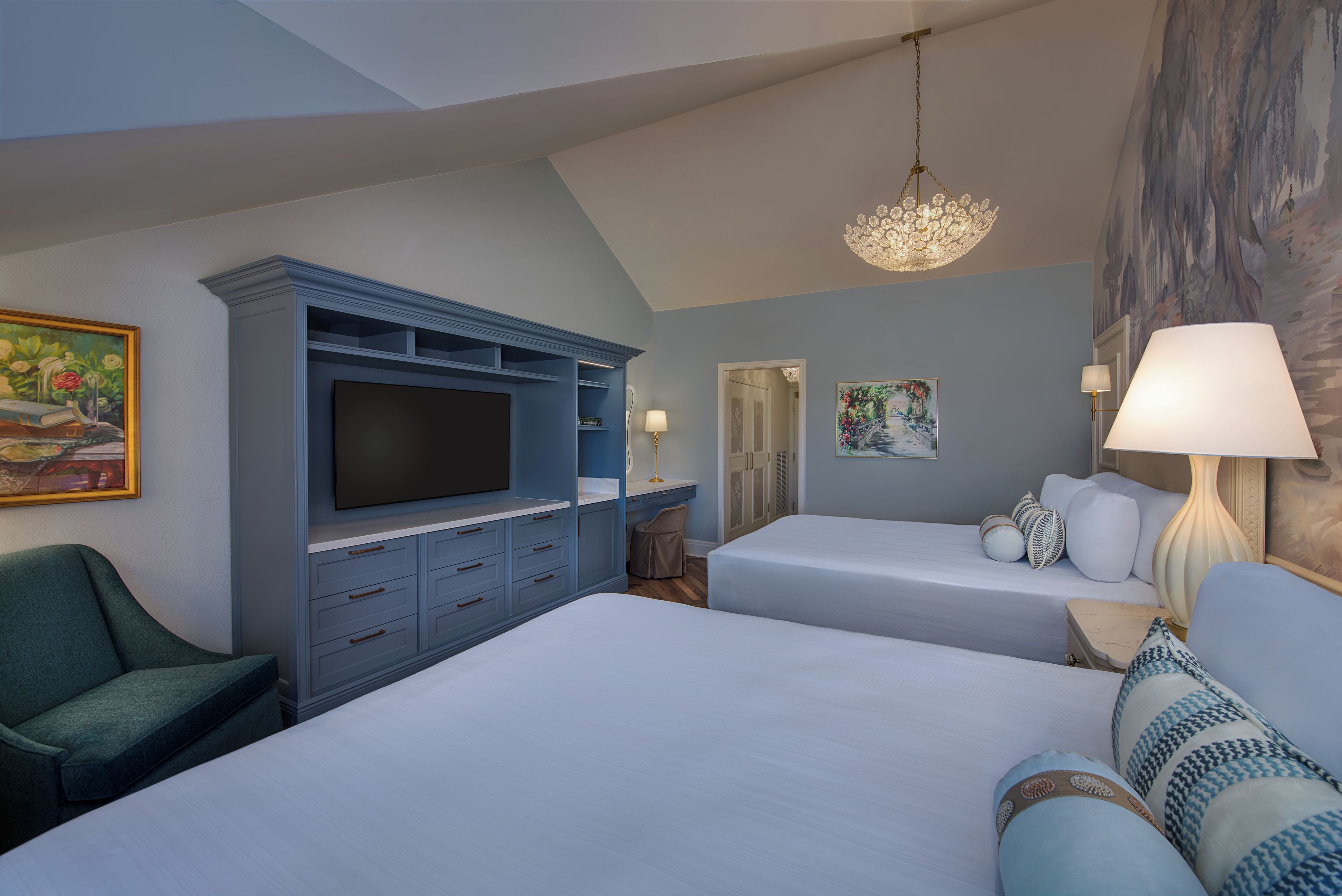 A hotel room with a large bed, a TV on a blue cabinet, a chair, two bedside tables with lamps, and artwork on the walls.