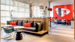 A modern lounge area with a sofa, tables, and chairs, featuring a large window and a red accent wall. - ibis Nuernberg City am Plaerrer (Nuremberg)