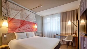 A hotel room with a bed, bedside tables, a desk, a chair, a large window with curtains, and a wall art piece. - ibis Nuernberg City am Plaerrer (Nuremberg)