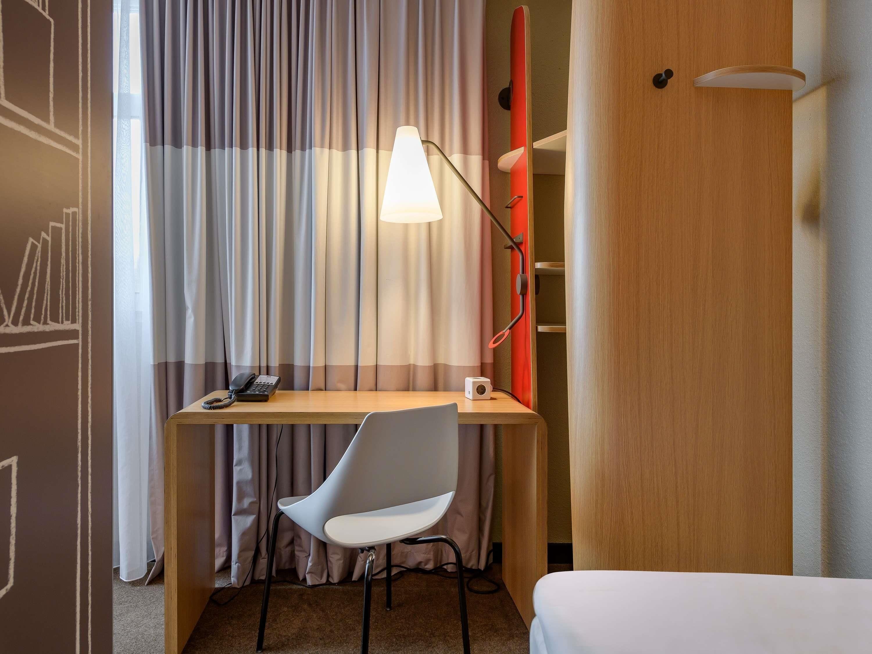 A hotel room with a wooden desk, a white chair, a telephone, a red object on the wall, and a bed.