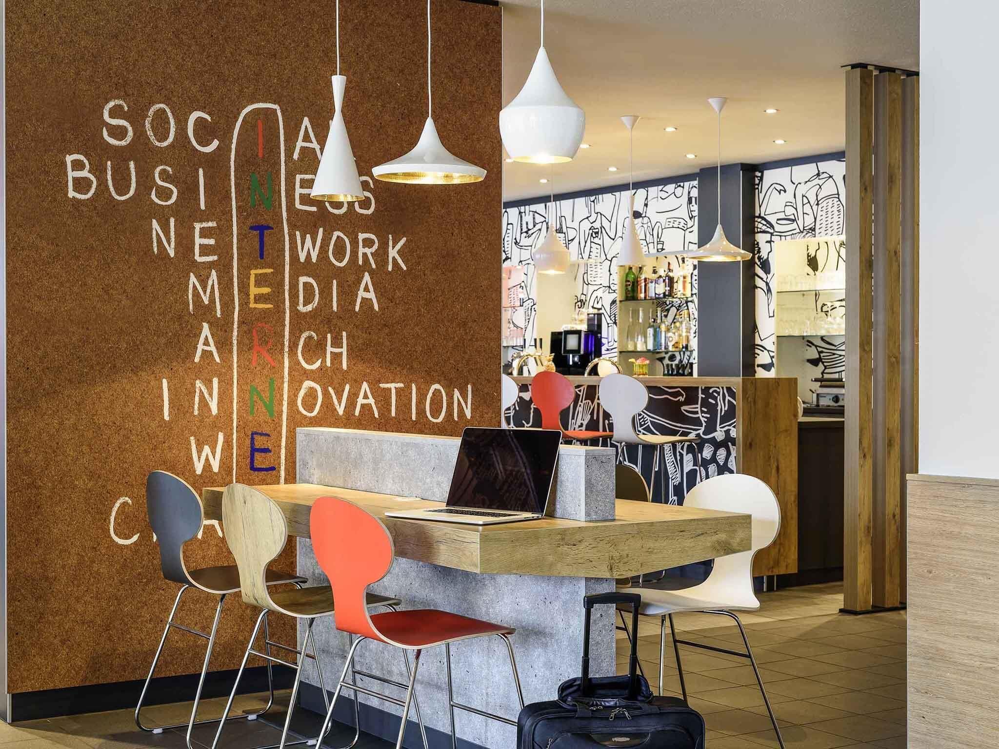 A modern workspace with a corkboard wall displaying business-related words, a wooden desk with a laptop, and colorful chairs.