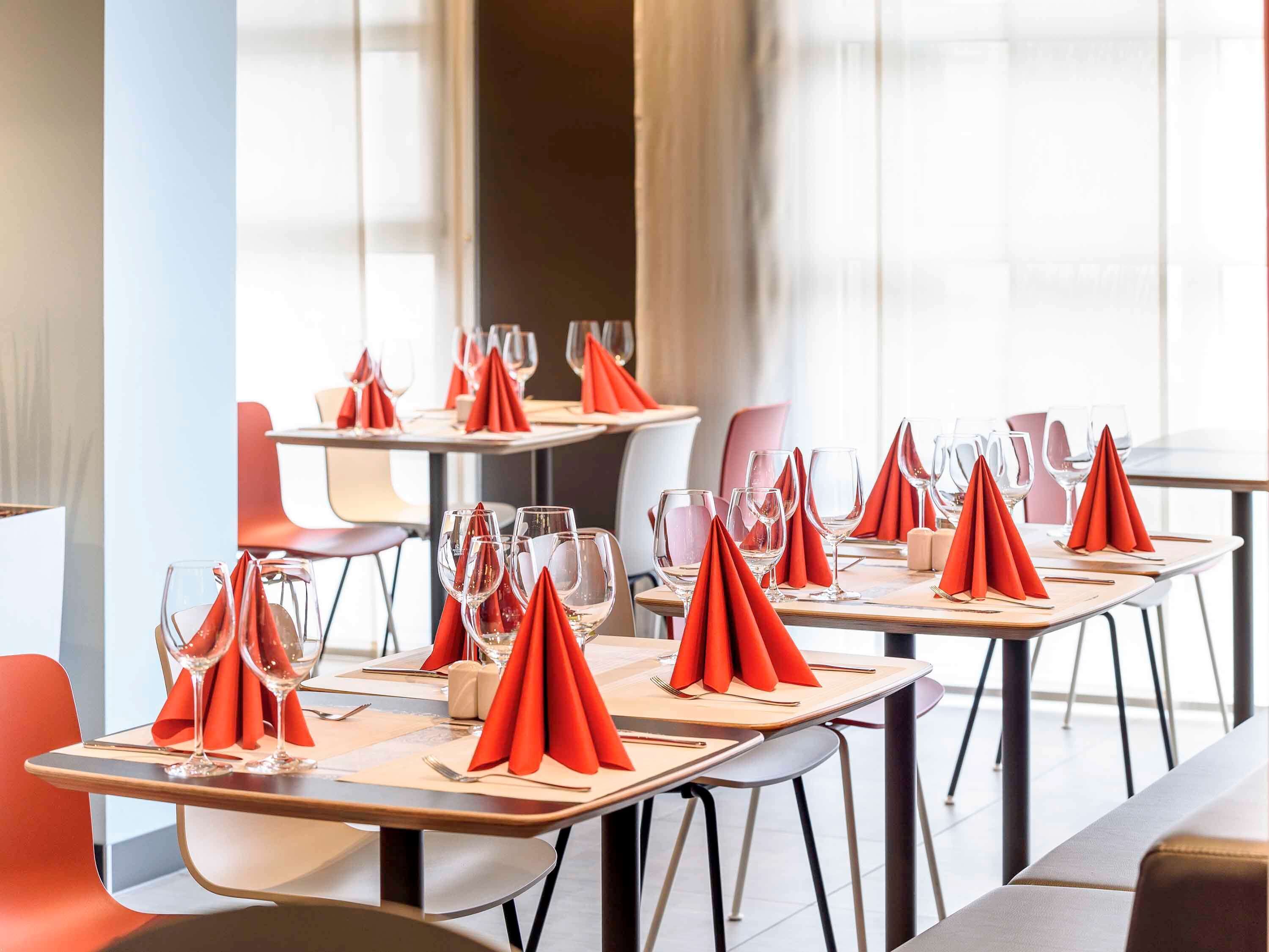 A restaurant with tables set with red napkins and wine glasses, modern chairs, and large windows.