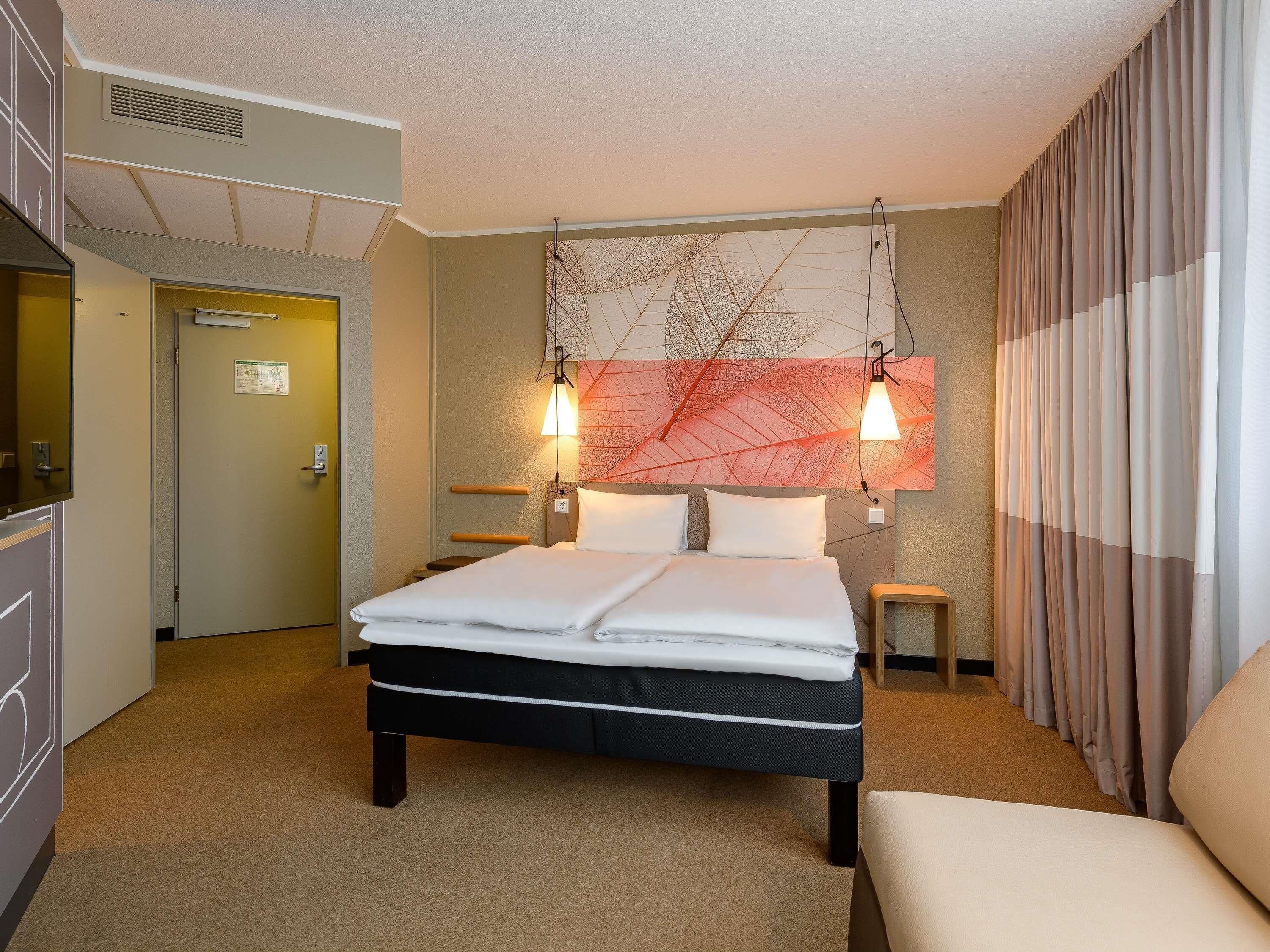 A hotel room with a bed, bedside tables, a headboard with a leaf pattern, a wall-mounted light fixture, and a curtain.