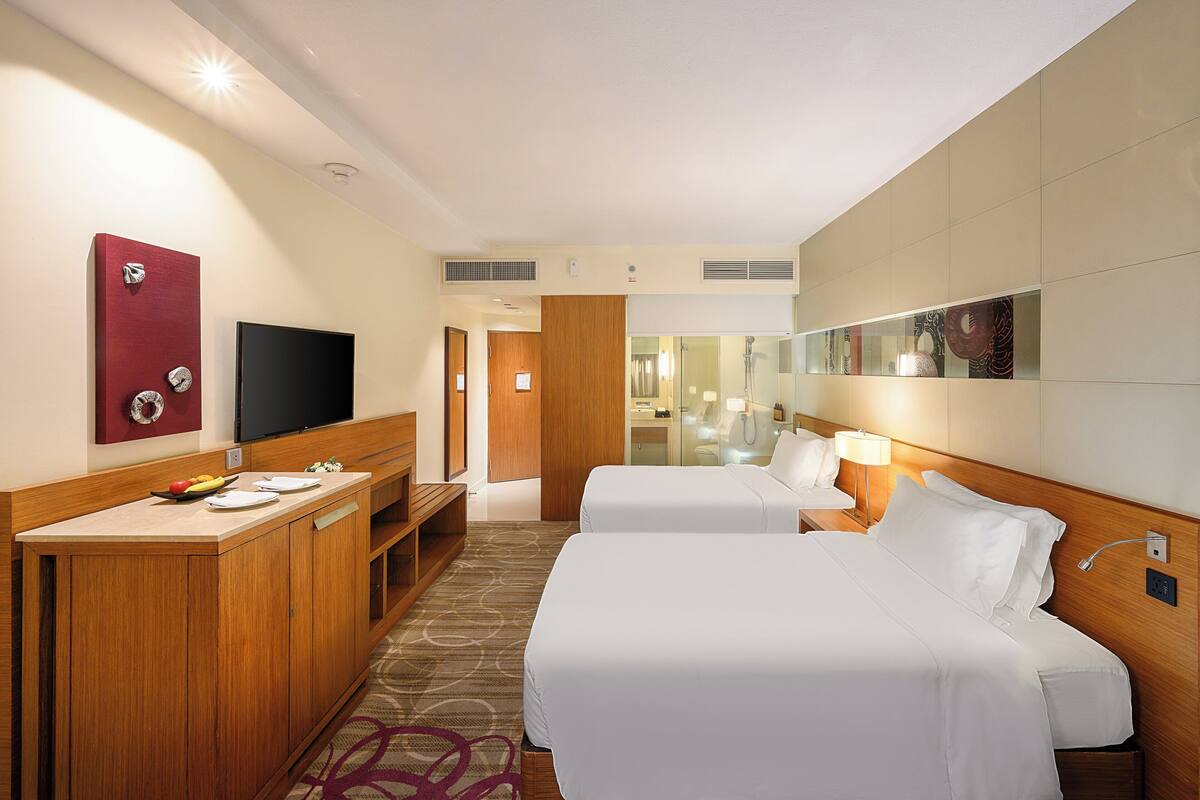 Ein Hotelzimmer mit zwei Betten, einem Schreibtisch, einem Fernseher und einem durch eine Glasscheibe einsehbaren Badezimmer.