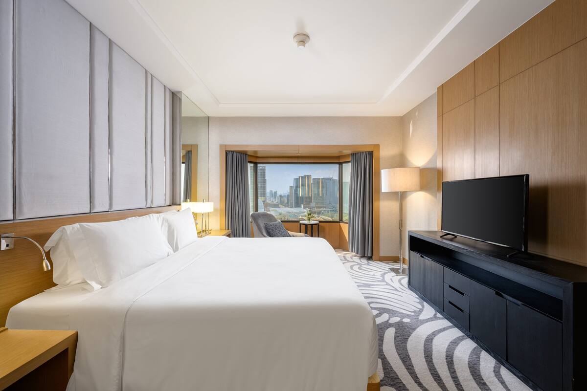Ein Hotelzimmer mit einem großen Bett, einem Flachbildfernseher, einem Schreibtisch mit Lampe, einem Sessel und Blick auf eine Stadtansicht durch ein Fenster.