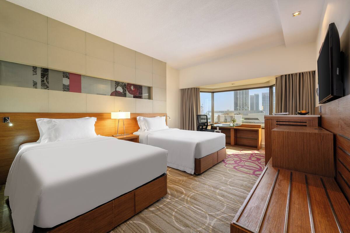 Ein Hotelzimmer mit zwei Betten, einem Schreibtisch, einem Fernseher und einem großen Fenster mit Blick auf die Stadt.