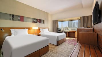 A hotel room with two beds, a desk, a TV, and a large window with a city view.