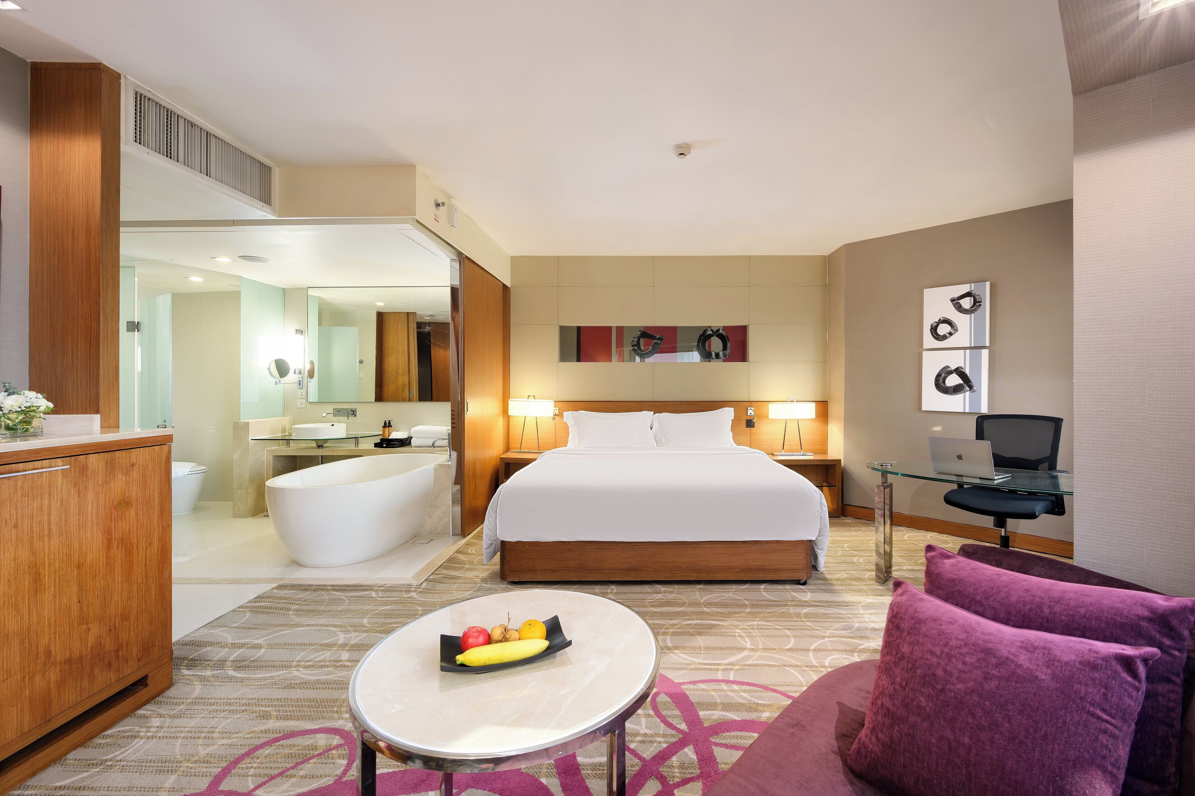 A hotel room with a bed, a desk with a laptop, a bathroom with a bathtub, and a dining table with fruit.