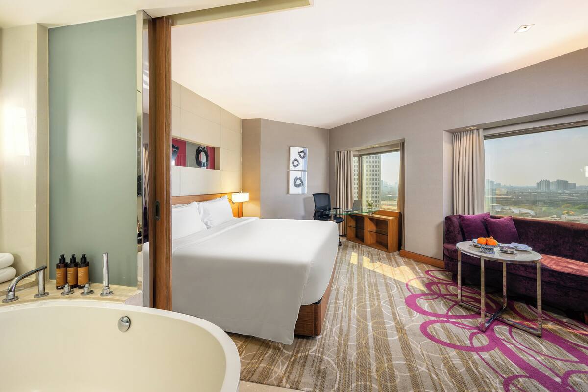 Ein Hotelzimmer mit einem großen Bett, einer Sitzecke mit Sofa und kleinem Tisch, einem Schreibtisch mit Stuhl, einem großen Fenster mit Stadtblick und einer Badewanne.