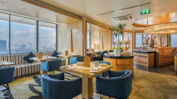 Un comedor moderno de hotel con vistas a la ciudad, con mesas y sillas dispuestas, y una zona de bar.